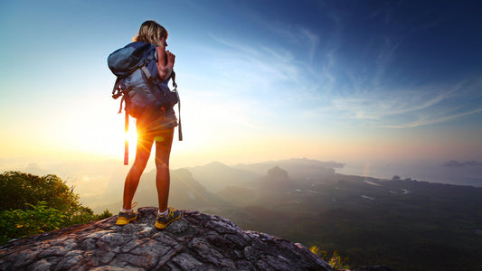 Young lady hiker standing with backpack on top of a mountain and enjoying sunrise